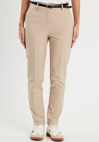 Pantalon beige ajusté avec une coupe slim, bordé de coutures discrètes, associé à une ceinture noire et des baskets blanches avec des accents beiges.