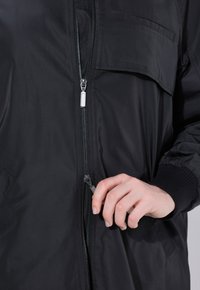 Chaqueta bomber negra con cierre frontal de cremallera, bolsillos laterales, puños elásticos y una textura lisa y mate. Incluye un bolsillo en el pecho.