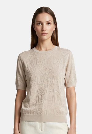 Frau mit glatten braunen Haaren, die ein beiges Kurzarm-Strickoberteil mit einem dezenten Blumenmuster und hellen Hosen trägt.