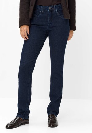 Person iført mørkeblå jeans med lige ben, sorte loafers, en mørk top og en mørkebrun jakke, stående mod en hvid baggrund.