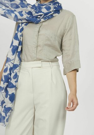 Femme portant une chemise beige à boutons avec des manches trois-quarts, un pantalon blanc taille haute et une écharpe à motifs bleus et blancs autour du cou.