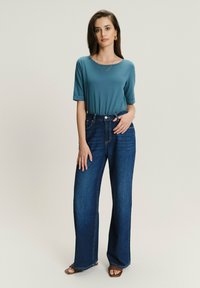 Zielona bawełniana koszulka z podwiniętymi rękawami, zestawiona z ciemnoniebieskimi dżinsami o wysokim stanie i szerokiej nogawce. Model nosi beżowe sandały oraz minimalną biżuterię.