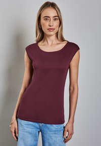 Burgundská bezrukávová halenka s širokým kulatým výstřihem, hladkým povrchem, přiléhavým střihem a rovním lemem, doplněná o světle modré džíny.