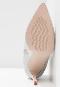 Chaussure stiletto blanche à bout pointu avec semelle beige texturée et talon fin, positionnée semelle vers le haut sur un fond blanc en coin.