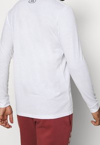 Biała koszula z długim rękawem wykonana z gładkiego materiału, z okrągłym dekoltem i logo z tyłu. Noszona z czerwonymi spodniami dresowymi.