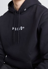 Felpa nera in cotone, con tasca a marsupio frontale e logo "VOLCOM" ricamato in bianco. Cordini regolabili con punte nere.