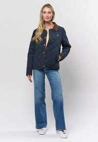 Vrouw die een marineblauwe doorgestikte jas met een bruine kraag draagt, een lichtbeige shirt, blauwe spijkerbroek en witte sneakers, staand tegen een effen achtergrond.