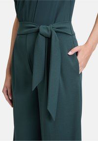 Pantaloni verdi a vita alta con una ampia cintura da annodare. Il materiale ha una texture liscia. Presenta tasche laterali e una vestibilità comoda.