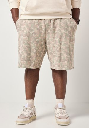 Person iført lyse beige shorts med camouflage-mønster, beige sneakers med hvide snørebånd og cremefarvede sokker, stående mod en ensfarvet baggrund.