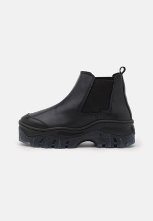 Botte Chelsea en cuir noir avec une semelle épaisse et robuste, des panneaux latéraux élastiques et une languette de traction sur un fond blanc uni.