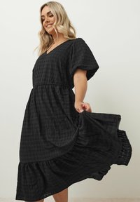 Musta midi-mekko laatikkomainen hihallinen, teksturoitu kangas, V-pääntie ja kerrostettu helma. Valmistettu kevyestä materiaalista, virtaava siluetti.