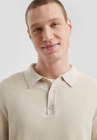 Beige gestricktes Poloshirt mit einem strukturierten Muster und einem kragen. Verfügt über drei Knöpfe an der Knopfleiste und einem leichten, weichen Stoff.