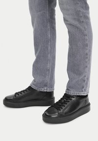 Čierne kožené tenisky s hladkým povrchom, okrúly špička, plochá podrážka a zladené čierne šnúrky, nosené s svetlo šedými džínami.