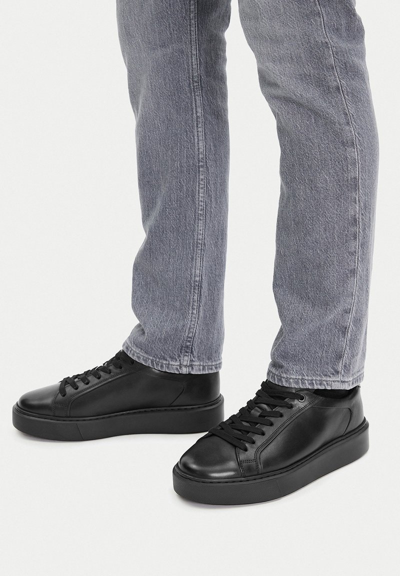 Čierne kožené tenisky s hladkým povrchom, okrúly špička, plochá podrážka a zladené čierne šnúrky, nosené s svetlo šedými džínami.