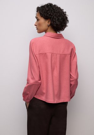 Blouse rose à manches longues en tissu doux. Comprend un col, un pli au dos et un ourlet arrondi. Associée à un pantalon foncé.