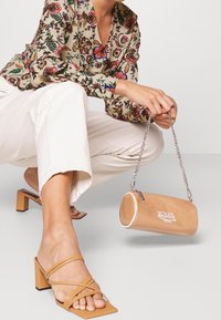 Tan färgad cylindrisk handväska med logotyp, kedjearm, och glansig yta. Ljusbeige blus med blommönster och krämfärgade byxor med vida ben.