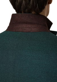 Ciemnozielona kurtka z wełny z domieszką, o teksturowanym wykończeniu, z kontrastowym brązowym filcowym kołnierzem. Widok z tyłu ukazuje kształt kołnierza i detale przeszycia.