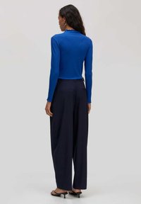 Enganliegende blaue Langarmbluse mit einem gewellten Ausschnitt, kombiniert mit locker sitzenden marineblauen Hosen und schwarzen Slippern.
