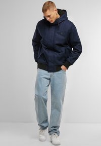 Jachetă cu glugă, de culoare bleumarin, cu închidere prin fermoar, buzunare laterale, manșete cu nervuri și interior moale. Asortată cu blugi largi, de culoare albastru deschis, și adidași albi.