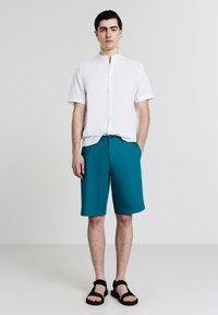 Camisetta bianca a maniche corte con colletto, pantaloni corti turchesi e sandali neri. La camicia presenta dei bottoni; i pantaloni corti hanno una texture liscia e morbida.