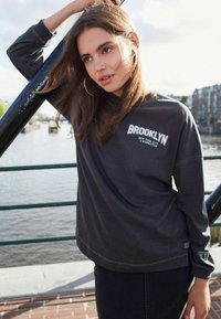 Harmaa pitkähihainen huppari, jossa on "BROOKLYN" painettuna rintaan, varustettuna ribatulla hihansuulla ja rennolla istuvuudella. Yhdistetty tummiin farkkuihin.