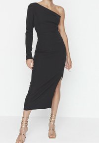 Robe midi noire, ajustée, avec un design asymétrique à une épaule. Elle présente une fente sur le côté et un accent en chaîne à l'épaule. Texture lisse.