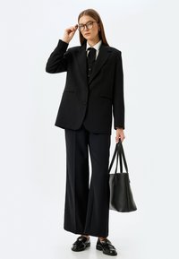 Schwarzer maßgeschneiderter Blazer über einem Hemd, kombiniert mit weit geschnittenen Hosen. Die Accessoires umfassen schwarze Loafers und eine strukturierte Tragetasche.
