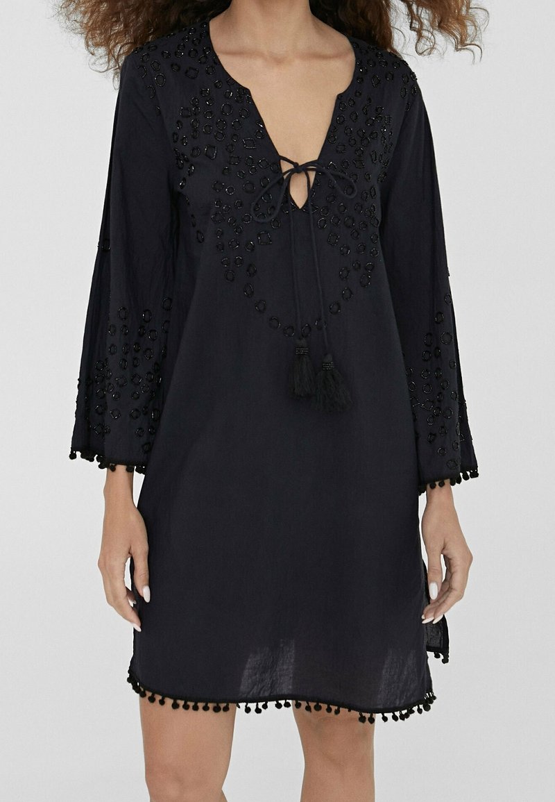Svart klänning med djup v-ringning, dekorativ pärlbroderi och luftiga ärmar. Har fransar vid halslinningen och en pom-pom dekoration vid fållen.