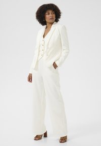 Blazer blanc ajusté avec des boutons dorés sur un gilet assorti et un pantalon à jambes larges. Tissu lisse, poches dans le pantalon, et chaussures à talons épais marron.
