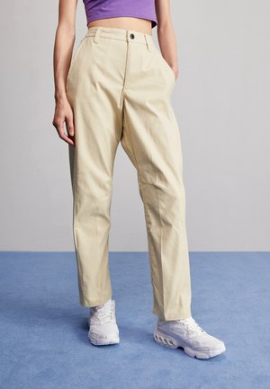 Person wearing beige high-waist pants, purple cropped top, and white athletic shoes standing on blue floor against gray wall.