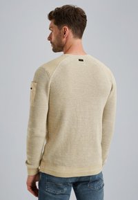 Beżowy sweter o fakturze, z prążkowanym dekoltem i mankietami, posiadający kieszeń na lewym rękawie oraz małą naszywkę z logo z tyłu.