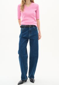 Kvinna i en kortärmad stickad topp i rosa, högmidjade blå jeans med ett svart bälte och svarta spetsiga skor mot en vit bakgrund.