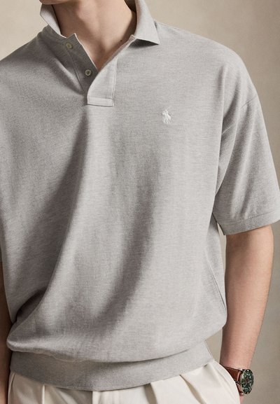 Άντρας που φοράει ανοιχτό γκρι πόλο με κοντά μανίκια και δύο κουμπιά, μικρό λευκό κεντημένο λογότυπο, μπεζ παντελόνι και σκοτεινό ρολόι χειρός.