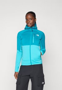 Giacca a vento turchese con cappuccio e chiusura a zip, caratterizzata da un design a blocchi di colore, due tasche laterali e logo sul petto. Tessuto di superficie liscia.