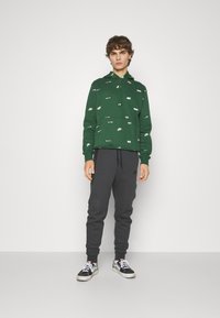 Grüne Kapuzenjacke mit weißen Logo-Drucken, kombiniert mit dunkelgrauen Jogginghosen. Das Modell trägt schwarze und weiße Turnschuhe. Einfarbiger Hintergrund.