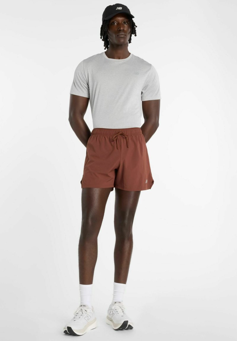 Grijs shirt met korte mouwen, bruine elastische korte broek met trekkoord, en witte sneakers. Model draagt een zwarte pet; minimale branding zichtbaar.