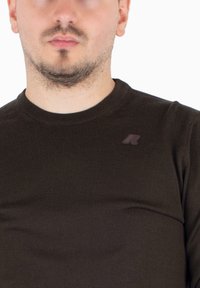 Pull en maille côtelée marron avec un col rond et un petit accent de marque sur la poitrine gauche. Texture douce et design ajusté.