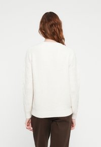 Cream sweater with a textured knit design, featuring cable-knit detailing on the sleeves and ribbed cuffs, worn with brown pants.