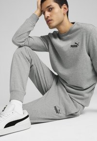 Grå sweatshirt og sweatpants, begge med sorte Puma-logoer. Hvide sneakers med sorte detaljer og teksturerede såler. Enkel, afslappet design.