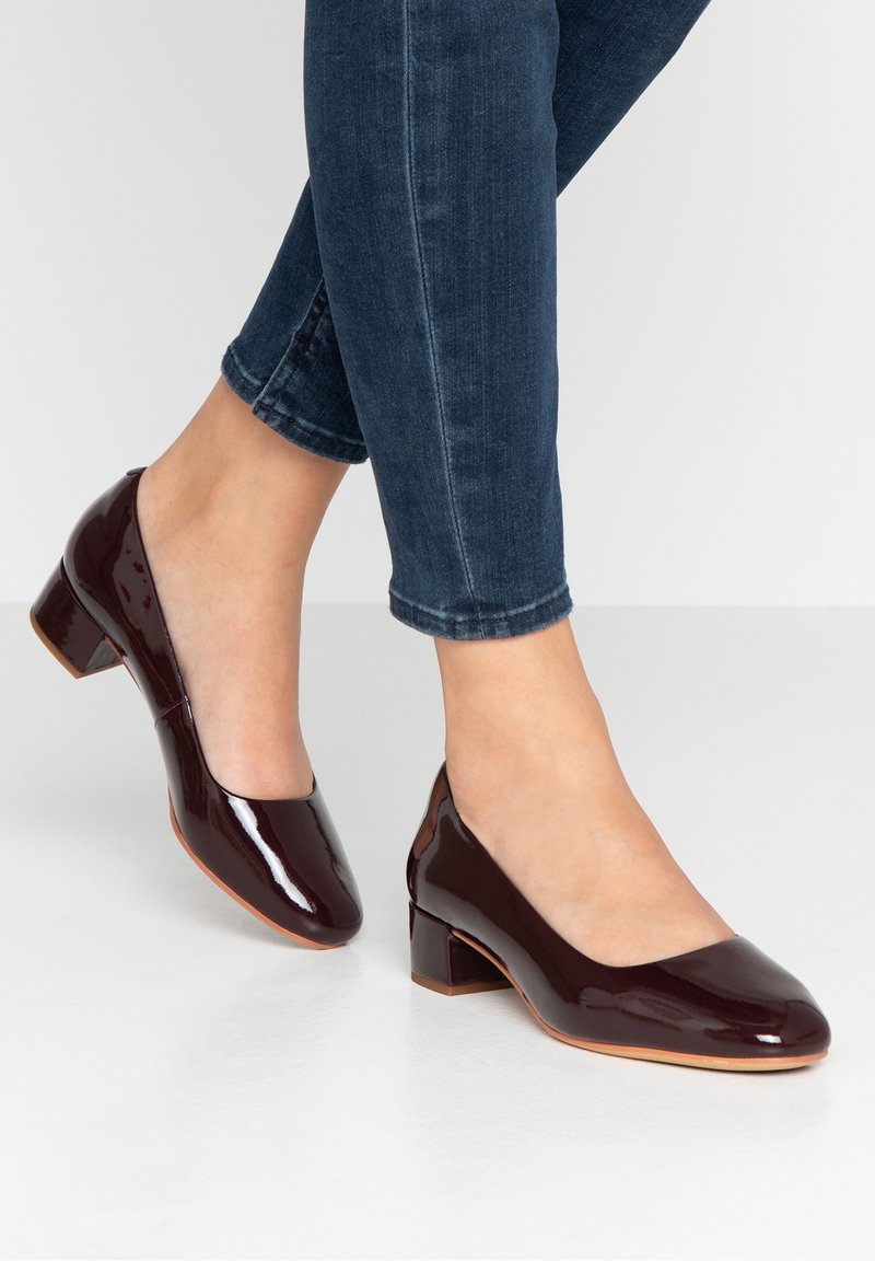 Chaussures à talon carré en cuir verni bordeaux avec un bout rond, un design épuré et une texture lisse, accompagnées de jeans en denim foncé.