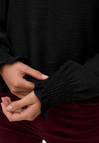 Mani che sistemano il polsino di una camicia nera a maniche lunghe con dettagli in elastico arricciato, abbinata a pantaloni in velluto a coste di un rosso scuro.