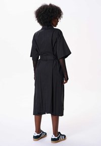 Czarna bawełniana sukienka midi z krótkim rękawem, z paskiem w talii i luźnym krojem. Noszona z zielono-białymi trampkami. Wyświetlony widok z tyłu.