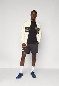 Lys kremfarge zip-jakke med marineblå stripe, svart T-skjorte, grå shorts, hvite sokker og marineblå sportsko med blå detaljer.