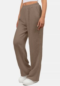 Hellbraune weit geschnittene Hose aus strukturiertem Stoff, mit einem elastischen Bund und seitlichen Taschen. Getragen mit weißen Sneakers.