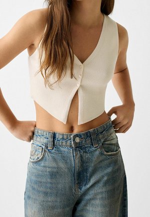 Frau trägt ein ärmelloses, beigefarbenes, geknöpftes Crop-Top und locker sitzende, ausgewaschene blaue Jeans, steht mit den Händen nahe der Taille.