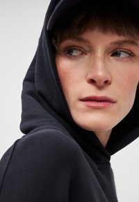 Čierna mikina s kapucňou, ktorá má mäkkú textúru a veľkú kapucňu. Tvár modelky je čiastočne viditeľná, s jemným make-upom a krátkymi vlasmi.