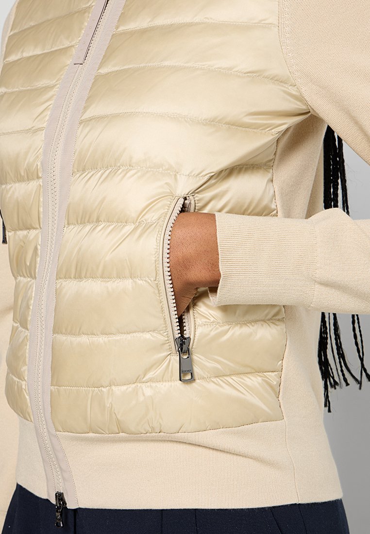 Helles beige Jackett mit einer gesteppten Pufferfront, glatten Strickärmel und einer Reißverschlusstasche mit schwarzen Hardware-Details.
