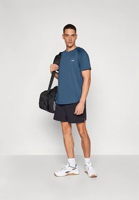 Férfi aktív ruházat: kék rövid ujjú ing, fekete rövidnadrág, fehér cipők barna részletekkel, fekete edzőtáska a kezében. Egyszerű kialakítás, könnyű anyag.