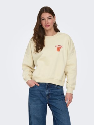 Vrouw met lang bruin haar, draagt gouden oorringen, een crèmekleurige sweatshirt met rode tekst "Out of Office" en een afbeelding van een drankje, en een blauwe spijkerbroek.
