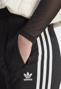 Pantalones deportivos negros con franjas laterales blancas, bolsillos con cremallera y logotipo de Adidas. Tejido texturizado y detalles ajustados en el puño.